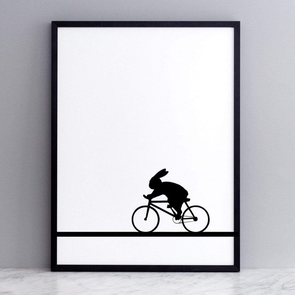 black and white image of HAM rabbit racing along on his bike.  Fun and playful series of prints.  Ideal for adults and children. Pictured here on marble surface with grey painted wall backdrop.